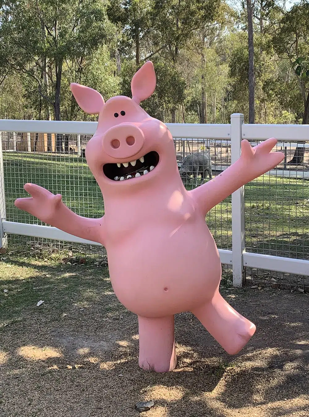Large, cheerful sculpture of a pink pig with arms outstretched, located in a park-like setting.