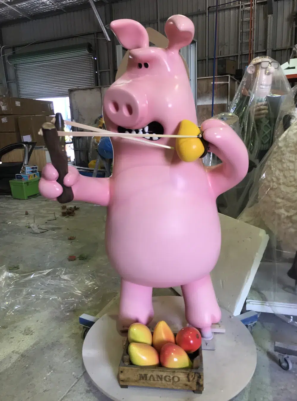 Bright pink animated pig sculpture playfully holding a slingshot and standing near a wooden crate of colorful mangoes in a workshop setting.