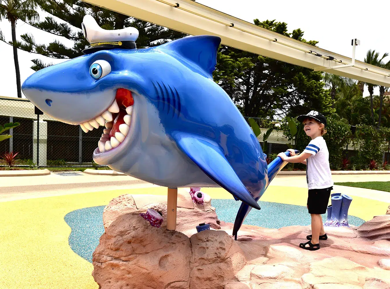 n outdoor play area with a child interacting with a large, playful blue shark sculpture adorned with a captain's hat; tall palm trees provide a backdrop.