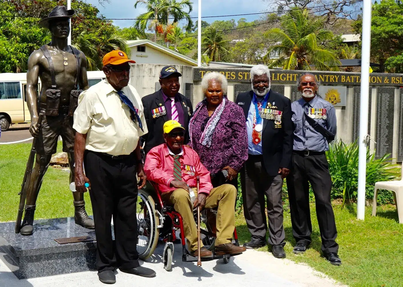Diverse group of individuals, some with military medals, posing proudly with a bronze soldier statue by Sculpt Studios in an outdoor setting.
